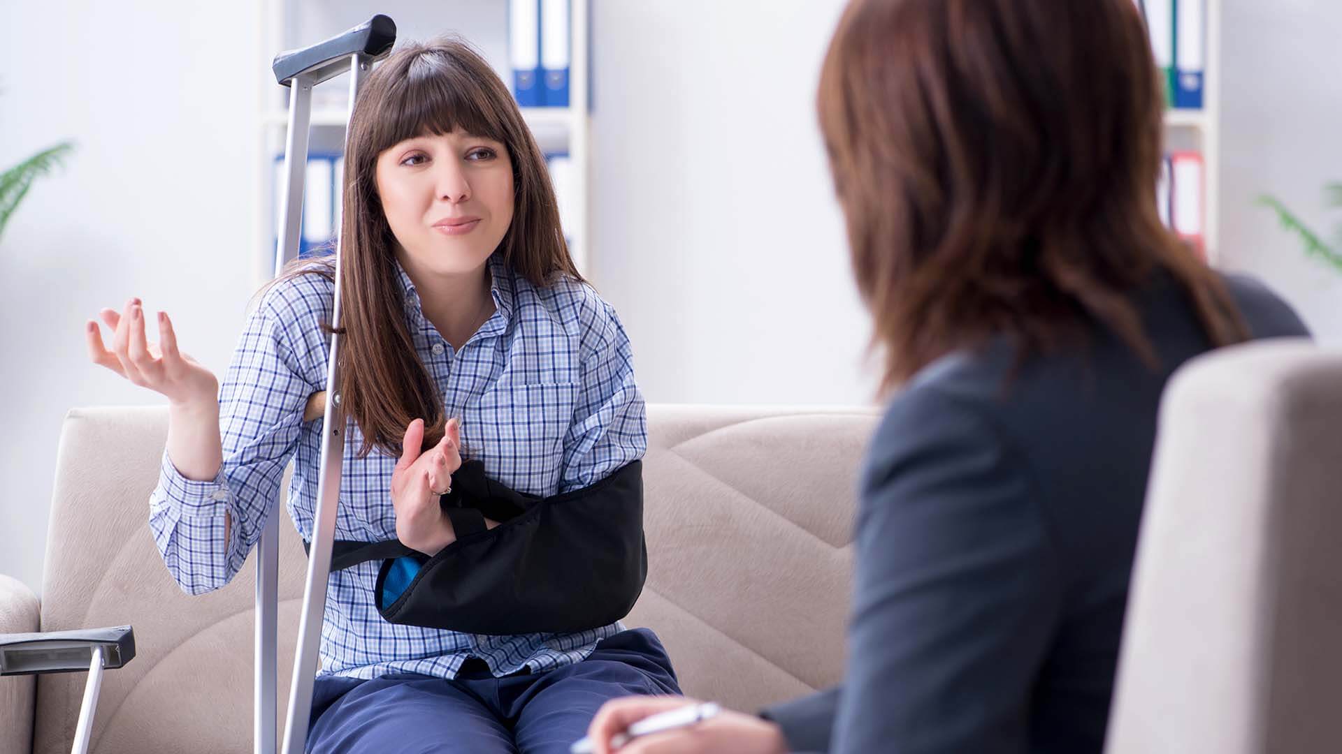 An injured woman on crutches explains her personal injury claim to a female lawyer or attorney as they decide if she should settle her case or go to court
